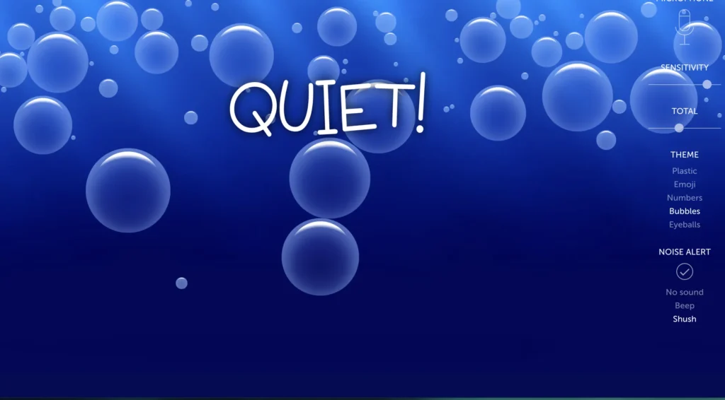 A classroom noise monitoring screen from BouncingBalls.org displaying large bubbles against a blue background. The text in the middle reads ‘QUIET!’ indicating a low noise level. A control panel on the right side shows options for sensitivity, total noise, and different themes such as plastic, emoji, numbers, and eyeballs